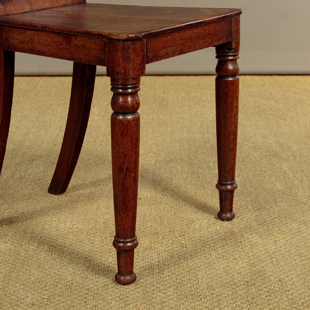 A pair of early nineteenth-century Regency mahogany hall chairs with carved oval backrests, scroll waist accents, solid wooden seats, and turned front legs. View VIII