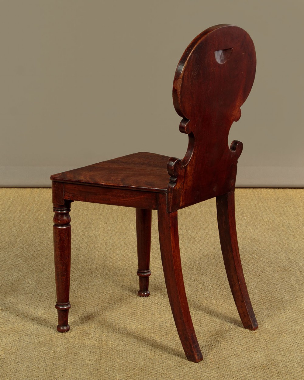A pair of early nineteenth-century Regency mahogany hall chairs with carved oval backrests, scroll waist accents, solid wooden seats, and turned front legs. View X