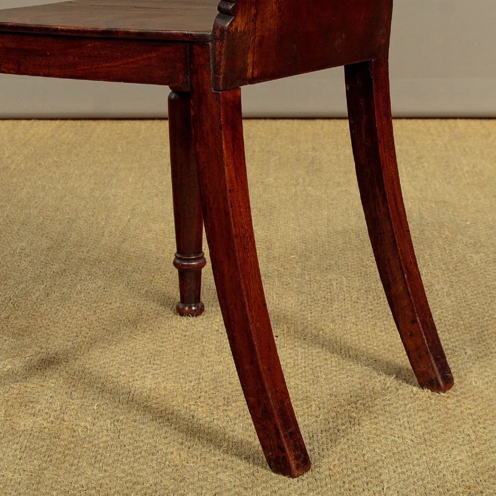 A pair of early nineteenth-century Regency mahogany hall chairs with carved oval backrests, scroll waist accents, solid wooden seats, and turned front legs. View XI
