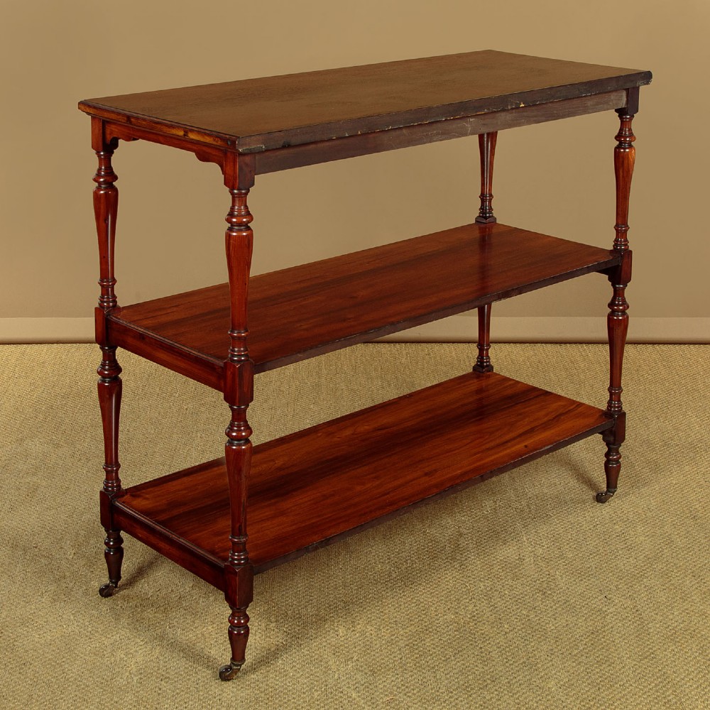 A three-tiered Victorian walnut dumb waiter featuring turned upright supports, open rectangular shelves, and brass castors.