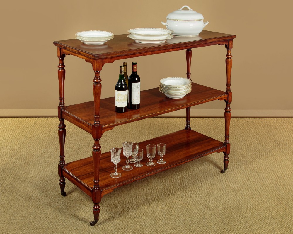A three-tiered Victorian walnut dumb waiter featuring turned upright supports, open rectangular shelves, and brass castors. View II