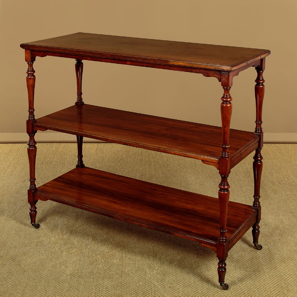 A three-tiered Victorian walnut dumb waiter featuring turned upright supports, open rectangular shelves, and brass castors. View III