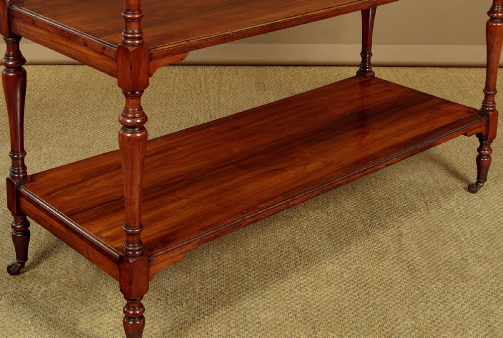 A three-tiered Victorian walnut dumb waiter featuring turned upright supports, open rectangular shelves, and brass castors. View VI