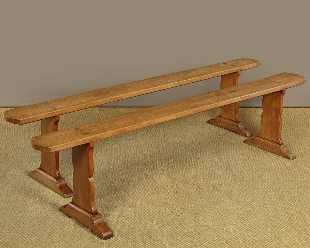A pair of long antique French cherrywood benches featuring simple plank seats supported by shaped trestle legs.
