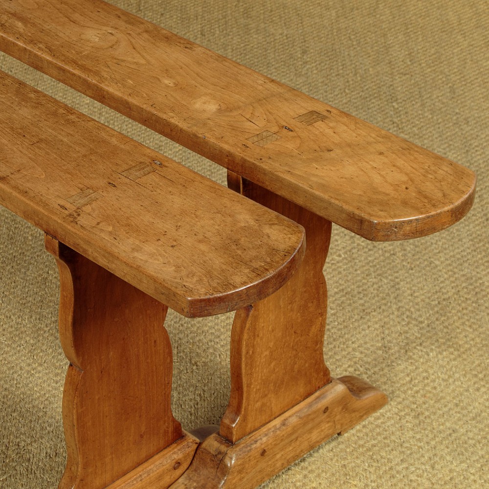 A pair of long antique French cherrywood benches featuring simple plank seats supported by shaped trestle legs. View III