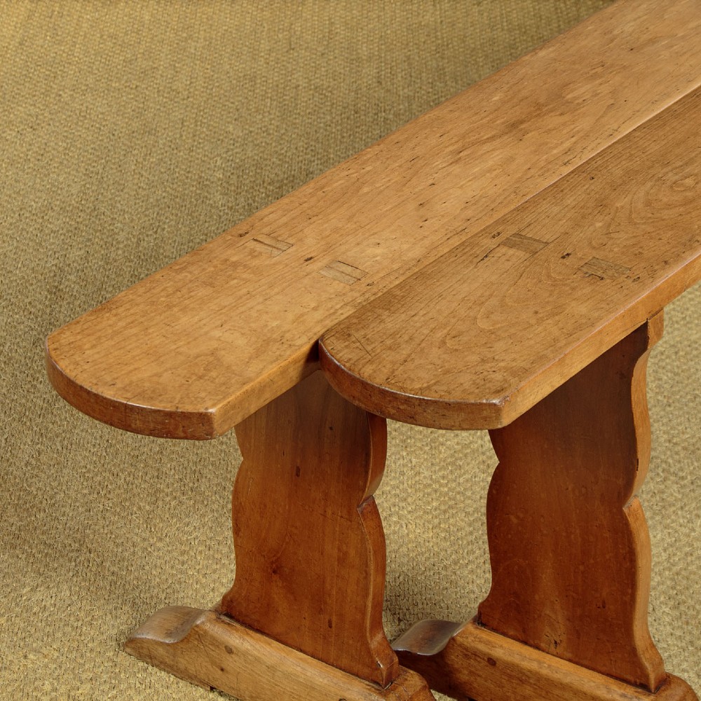 A pair of long antique French cherrywood benches featuring simple plank seats supported by shaped trestle legs. View IV