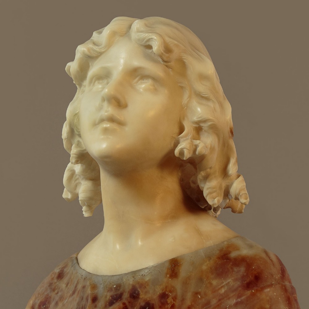 A carved alabaster bust depicting a young woman with shoulder-length wavy hair, wearing a dark amber stone dress on a matching plinth base. View V