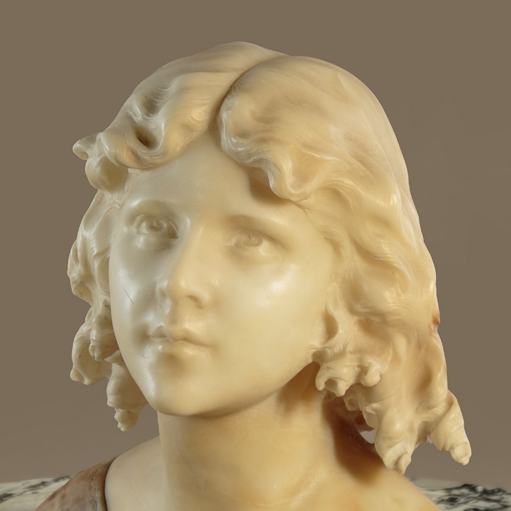A carved alabaster bust depicting a young woman with shoulder-length wavy hair, wearing a dark amber stone dress on a matching plinth base. View VI