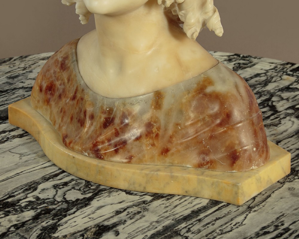 A carved alabaster bust depicting a young woman with shoulder-length wavy hair, wearing a dark amber stone dress on a matching plinth base. View IX