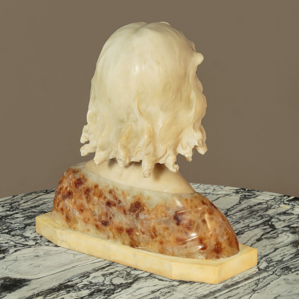A carved alabaster bust depicting a young woman with shoulder-length wavy hair, wearing a dark amber stone dress on a matching plinth base. View X