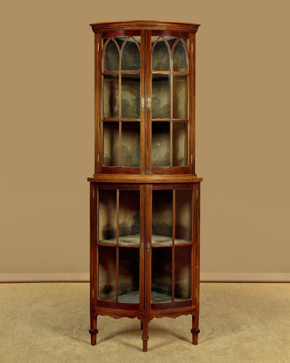 A two-tiered mahogany bow front corner display cabinet featuring upper gothic arch astragal glazing and lower glass panels.