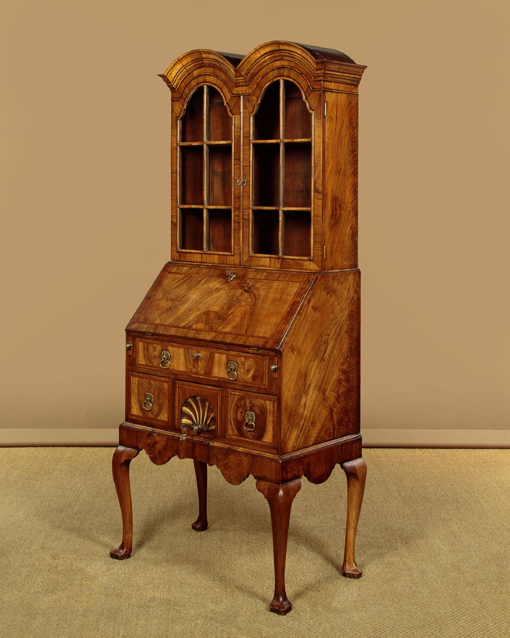 A Queen Anne style walnut bureau bookcase featuring a double-domed top, glazed upper doors, a slant-front writing slope, lower drawers with a carved shell motif, and slender cabriole legs.