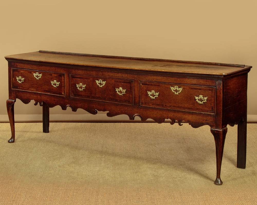 An eighteenth-century dark oak dresser base featuring three drawers with brass handles, a scalloped apron, and front cabriole legs.