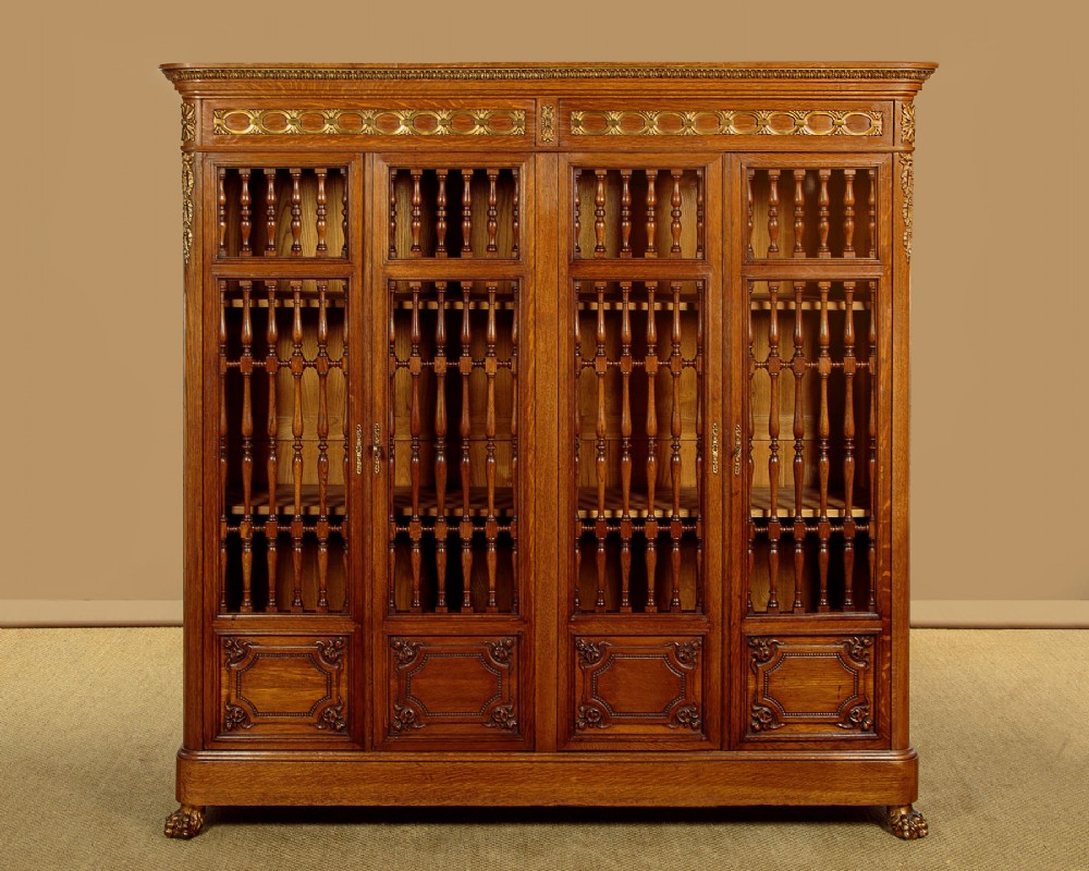 A large four-door oak bookcase featuring decorative turned spindles on the upper door sections, gilded brass frieze mounts, and carved lower timber panels.