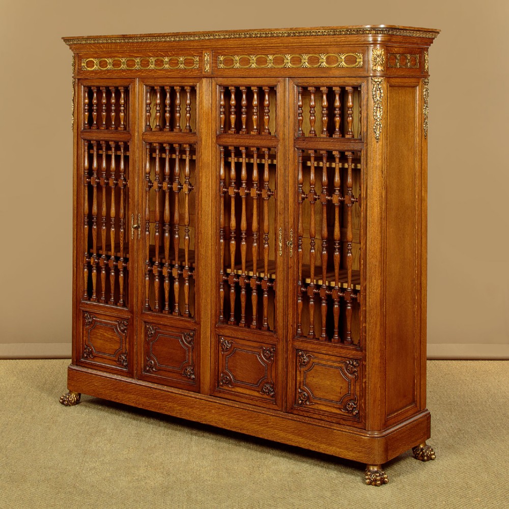 A large four-door oak bookcase featuring decorative turned spindles on the upper door sections, gilded brass frieze mounts, and carved lower timber panels. View III
