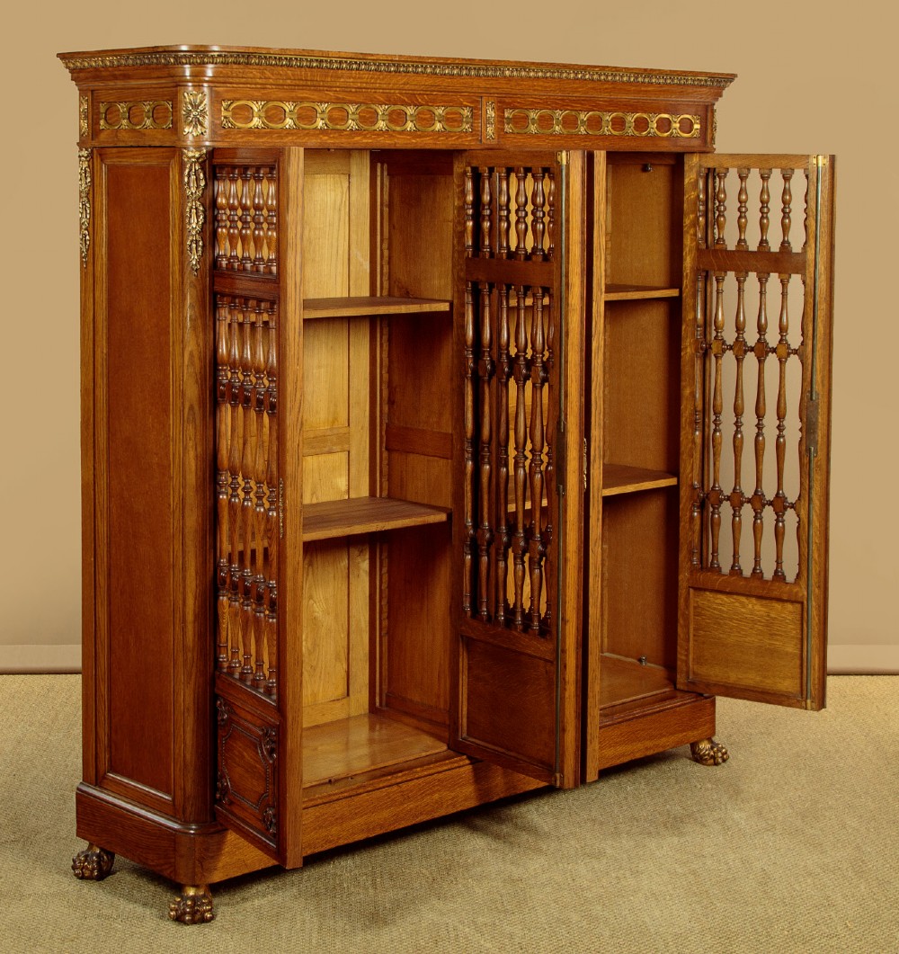 A large four-door oak bookcase featuring decorative turned spindles on the upper door sections, gilded brass frieze mounts, and carved lower timber panels. View IV