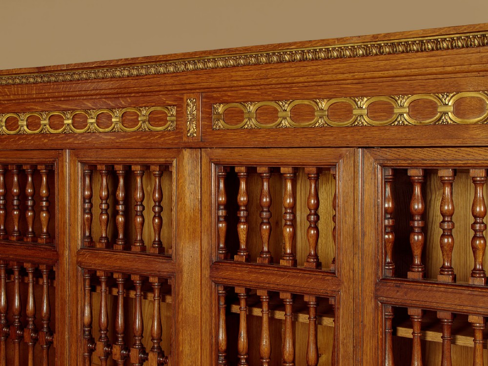 A large four-door oak bookcase featuring decorative turned spindles on the upper door sections, gilded brass frieze mounts, and carved lower timber panels. View V