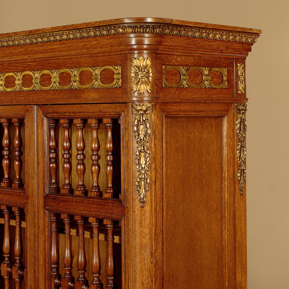 A large four-door oak bookcase featuring decorative turned spindles on the upper door sections, gilded brass frieze mounts, and carved lower timber panels. View VI