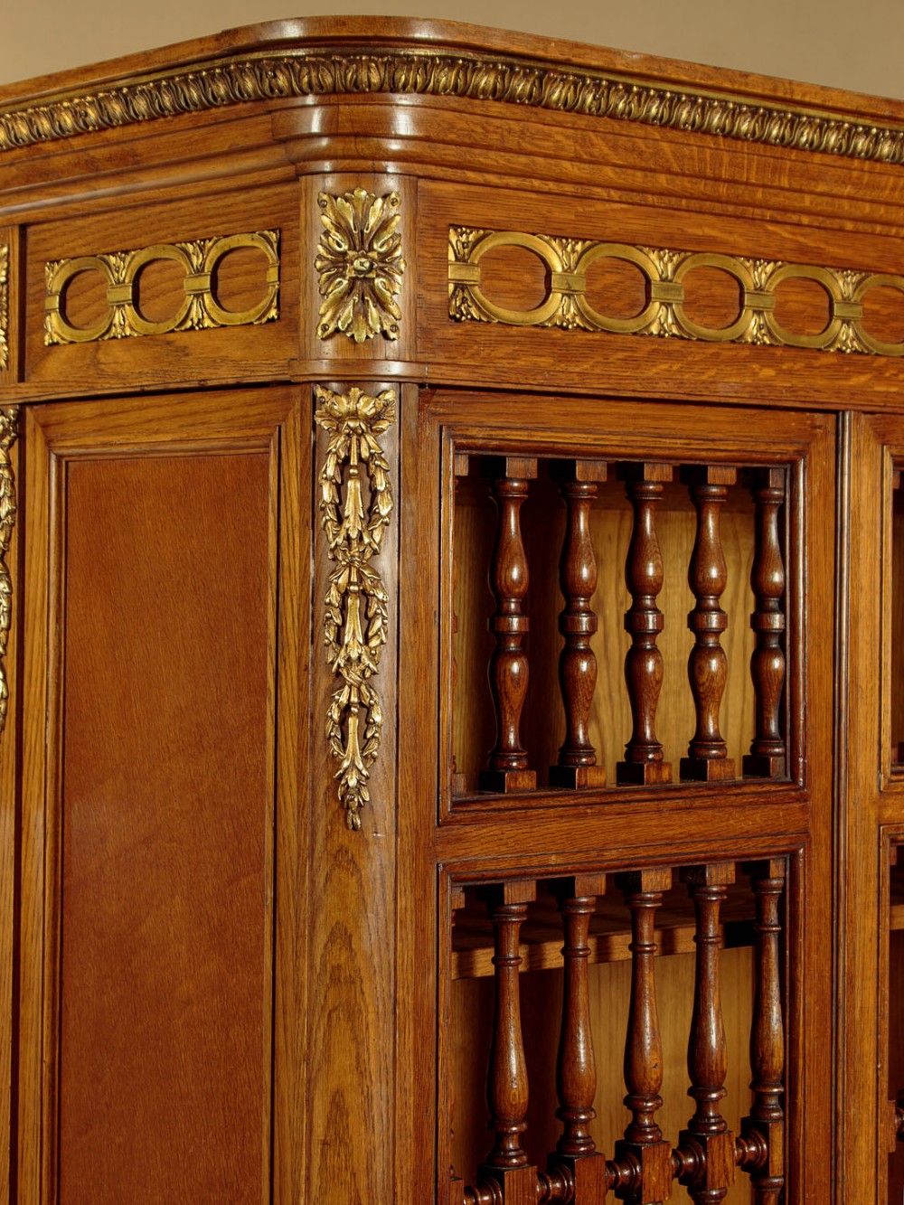 A large four-door oak bookcase featuring decorative turned spindles on the upper door sections, gilded brass frieze mounts, and carved lower timber panels. View VII