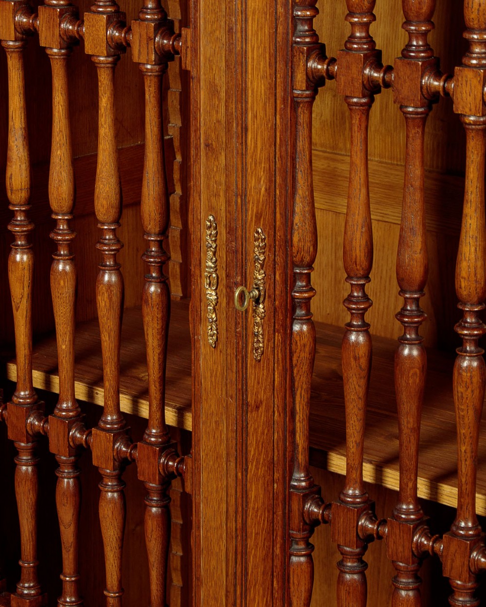 A large four-door oak bookcase featuring decorative turned spindles on the upper door sections, gilded brass frieze mounts, and carved lower timber panels. View VIII