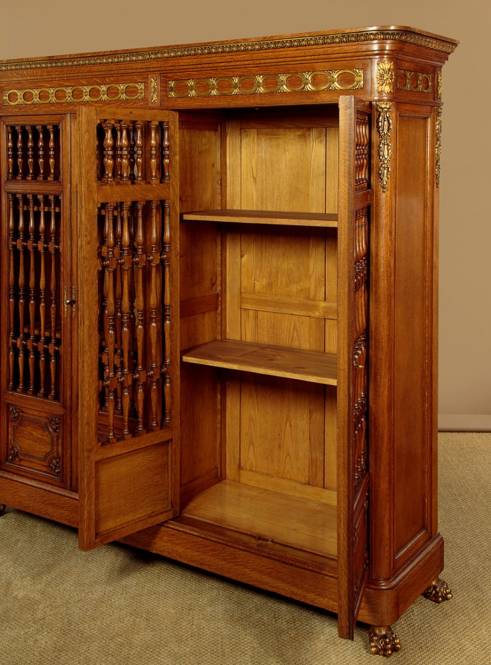 A large four-door oak bookcase featuring decorative turned spindles on the upper door sections, gilded brass frieze mounts, and carved lower timber panels. View IX