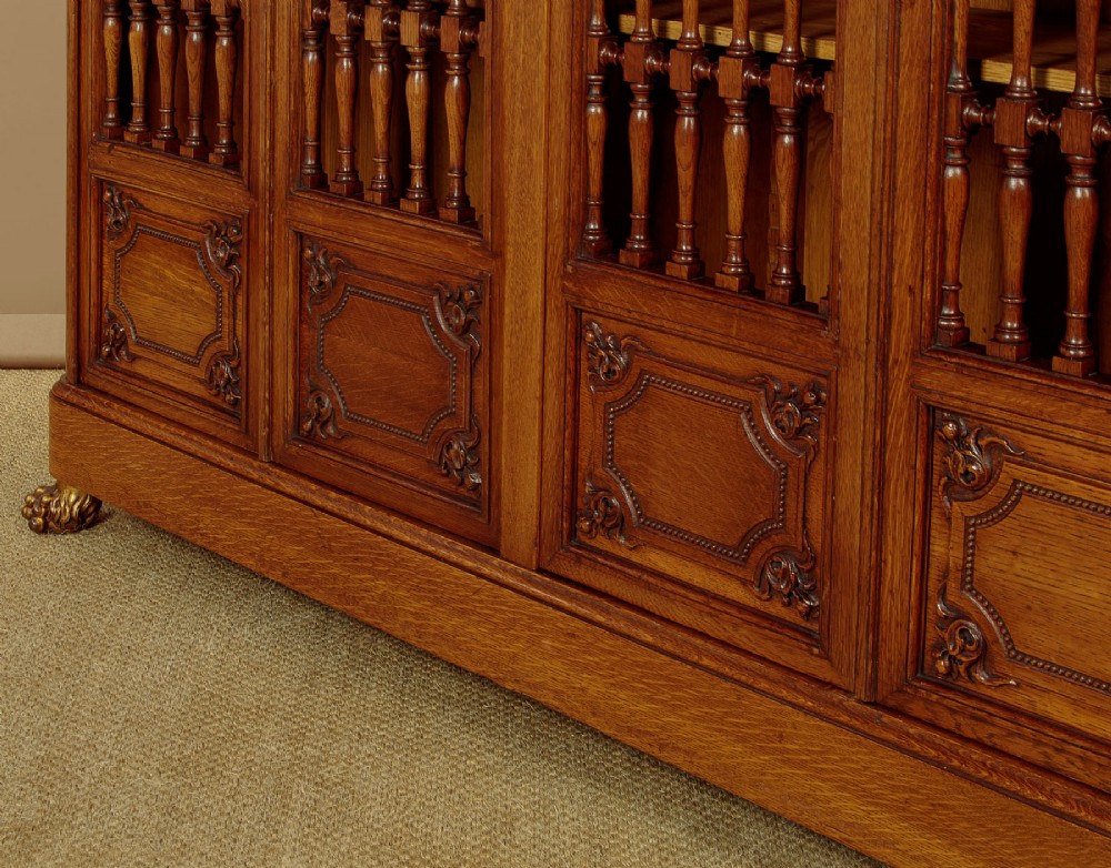 A large four-door oak bookcase featuring decorative turned spindles on the upper door sections, gilded brass frieze mounts, and carved lower timber panels. View X