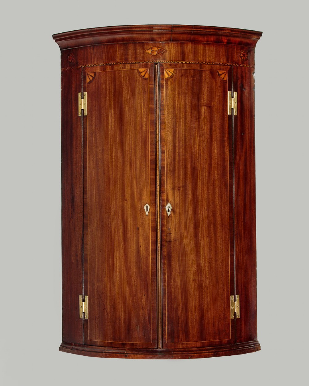 A George III bow-front mahogany corner cupboard featuring twin doors with inlaid fan motifs, crossbanded borders, and brass H-hinges.