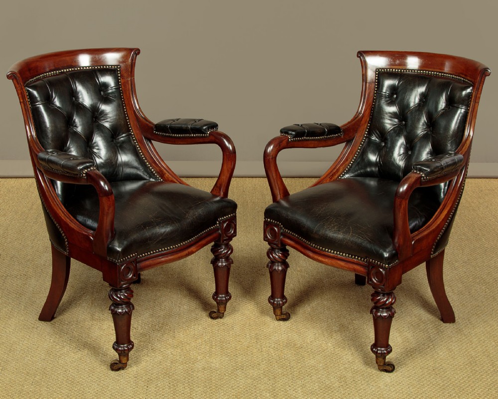 A pair of William IV mahogany library chairs featuring deep-buttoned dark leather upholstery, padded armrests, brass stud trim, turned carved front legs, and castors.