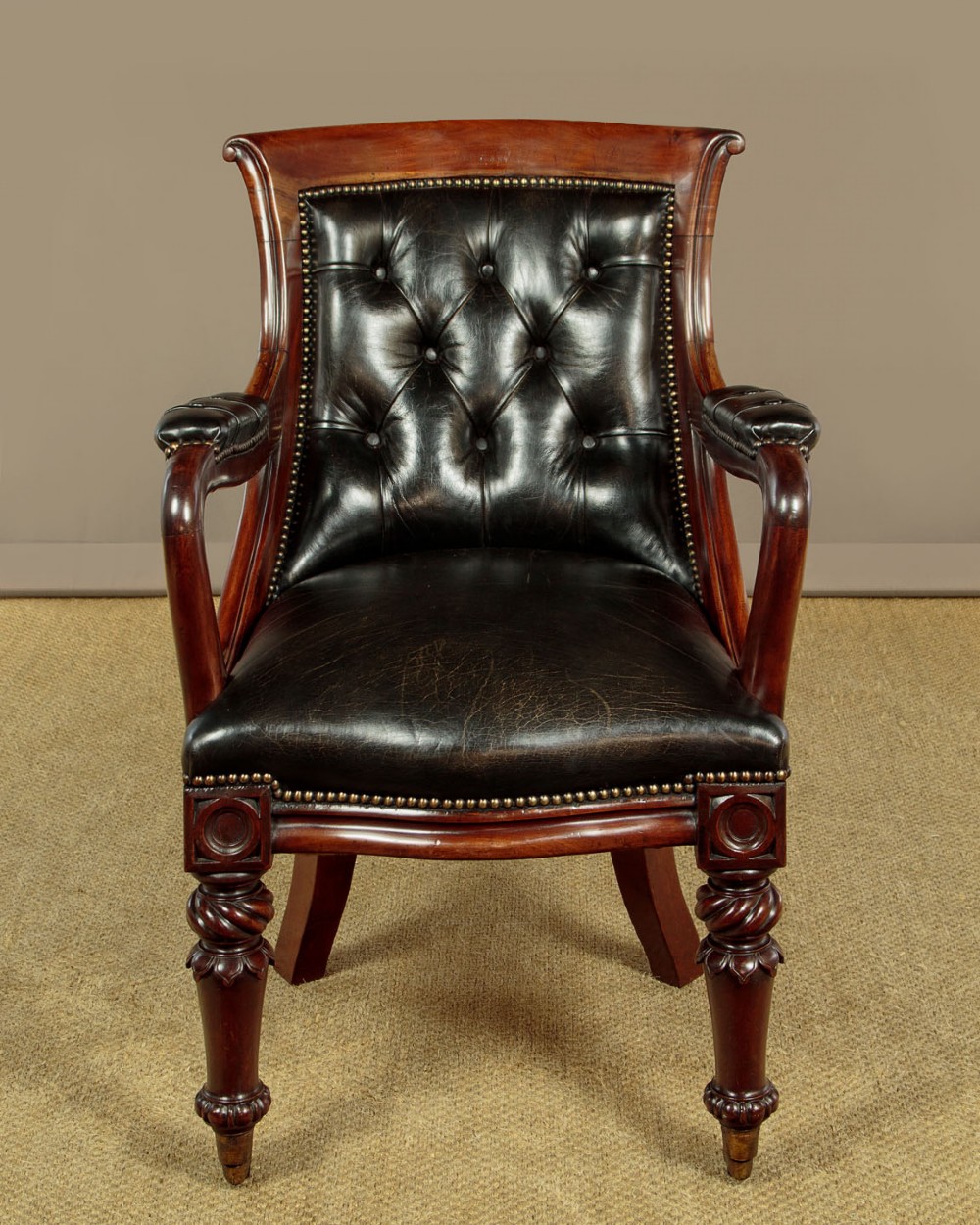 A pair of William IV mahogany library chairs featuring deep-buttoned dark leather upholstery, padded armrests, brass stud trim, turned carved front legs, and castors. View IV