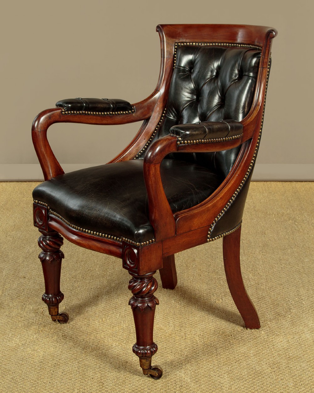 A pair of William IV mahogany library chairs featuring deep-buttoned dark leather upholstery, padded armrests, brass stud trim, turned carved front legs, and castors. View V