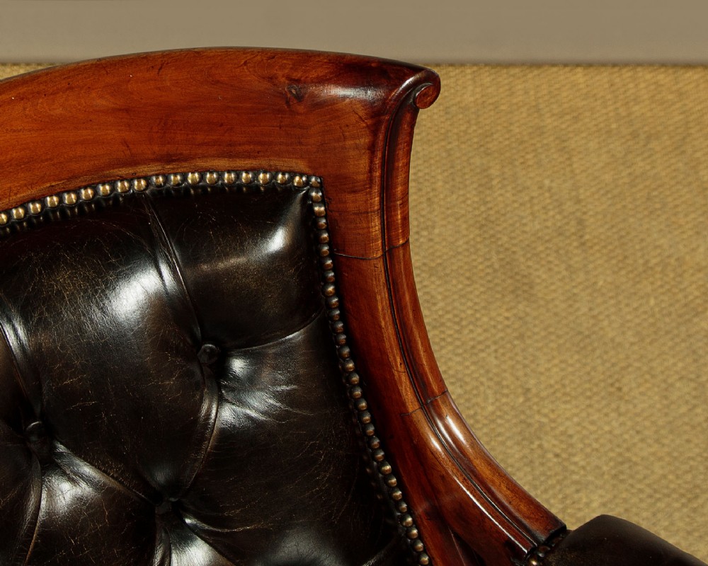 A pair of William IV mahogany library chairs featuring deep-buttoned dark leather upholstery, padded armrests, brass stud trim, turned carved front legs, and castors. View VI