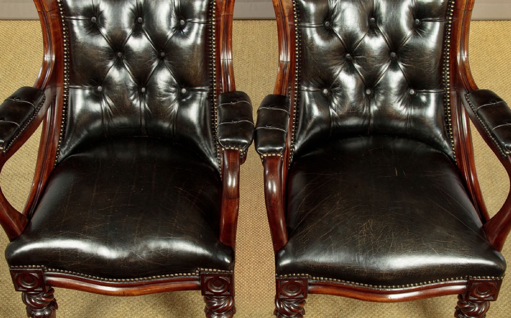A pair of William IV mahogany library chairs featuring deep-buttoned dark leather upholstery, padded armrests, brass stud trim, turned carved front legs, and castors. View IX