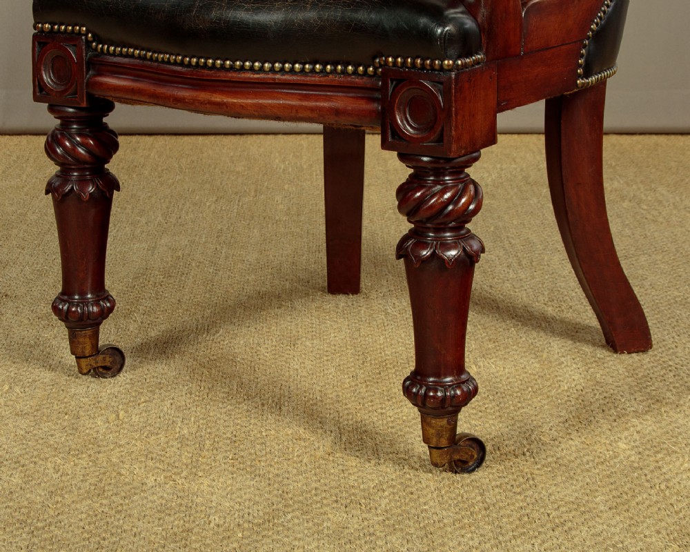 A pair of William IV mahogany library chairs featuring deep-buttoned dark leather upholstery, padded armrests, brass stud trim, turned carved front legs, and castors. View X
