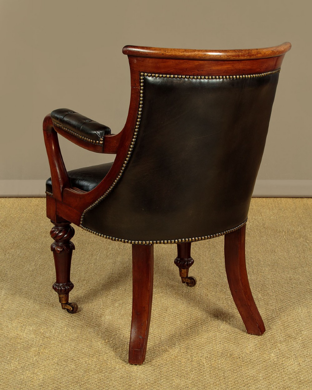 A pair of William IV mahogany library chairs featuring deep-buttoned dark leather upholstery, padded armrests, brass stud trim, turned carved front legs, and castors. View XII