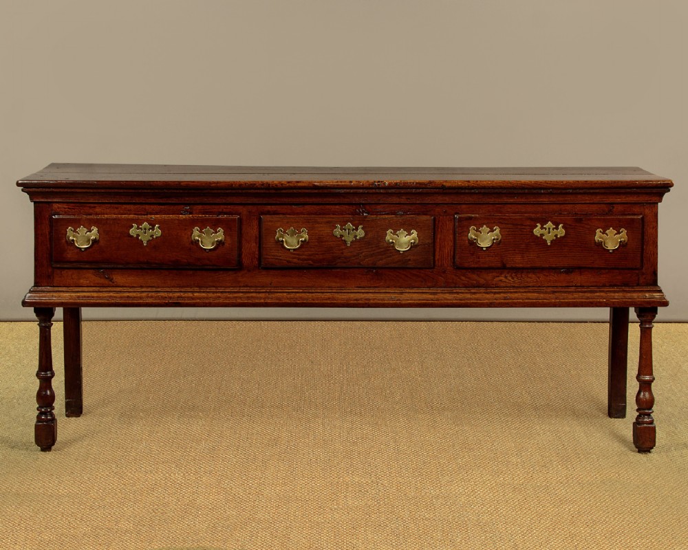 An 18th-century dark oak dresser base featuring three inline drawers with decorative brass drop handles and turned front legs.
