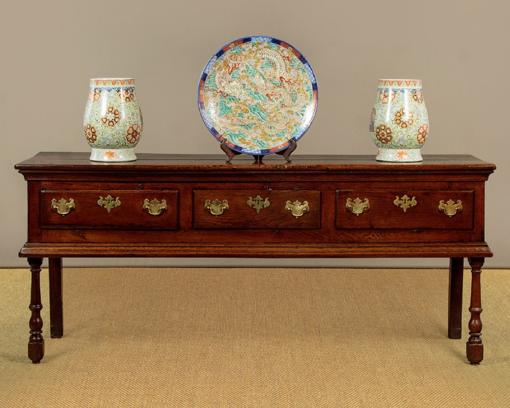 An 18th-century dark oak dresser base featuring three inline drawers with decorative brass drop handles and turned front legs. View II