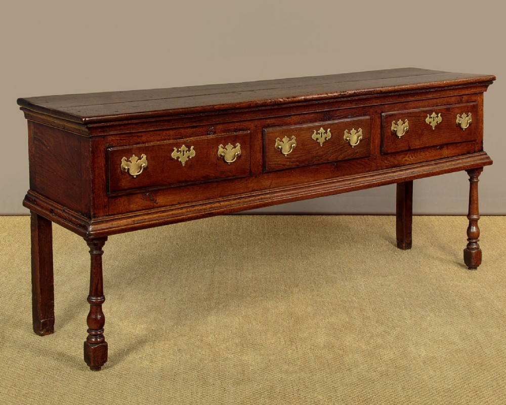 An 18th-century dark oak dresser base featuring three inline drawers with decorative brass drop handles and turned front legs. View III