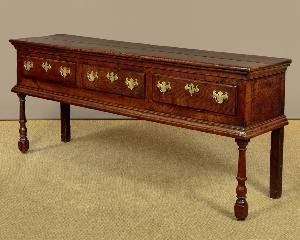 An 18th-century dark oak dresser base featuring three inline drawers with decorative brass drop handles and turned front legs. View IV