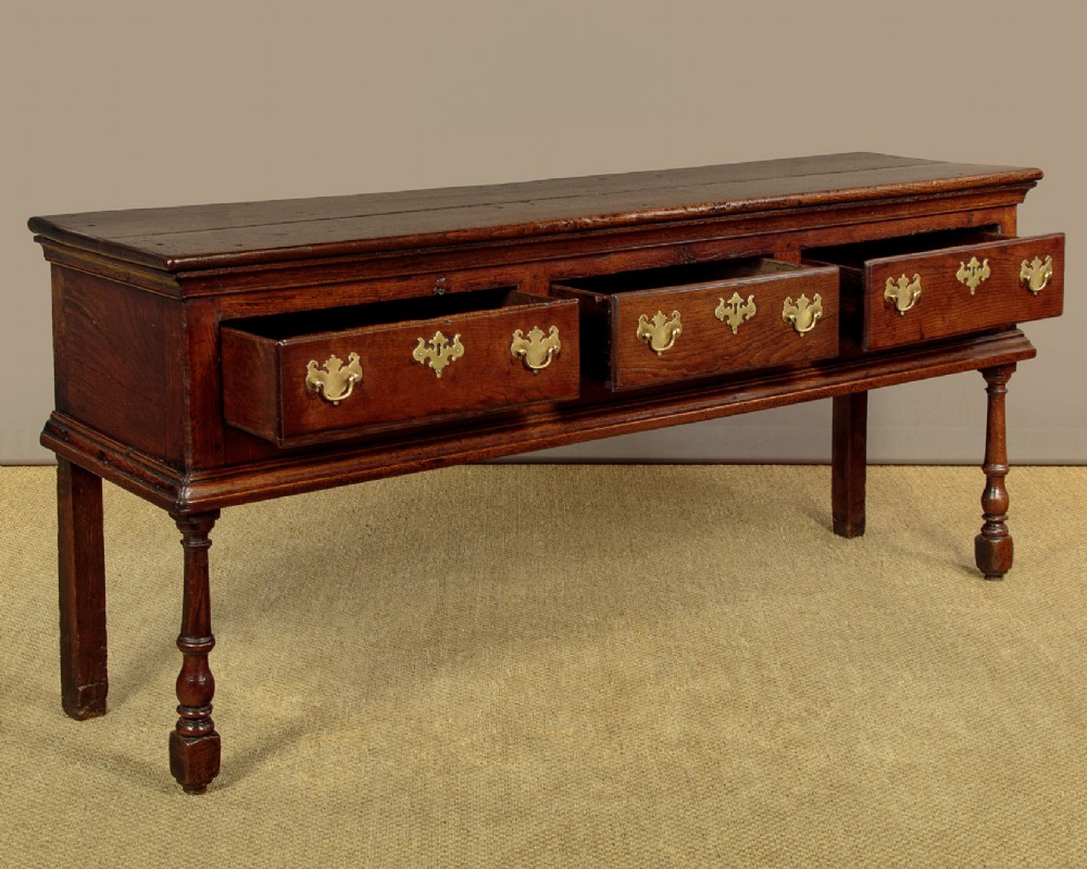 An 18th-century dark oak dresser base featuring three inline drawers with decorative brass drop handles and turned front legs. View V