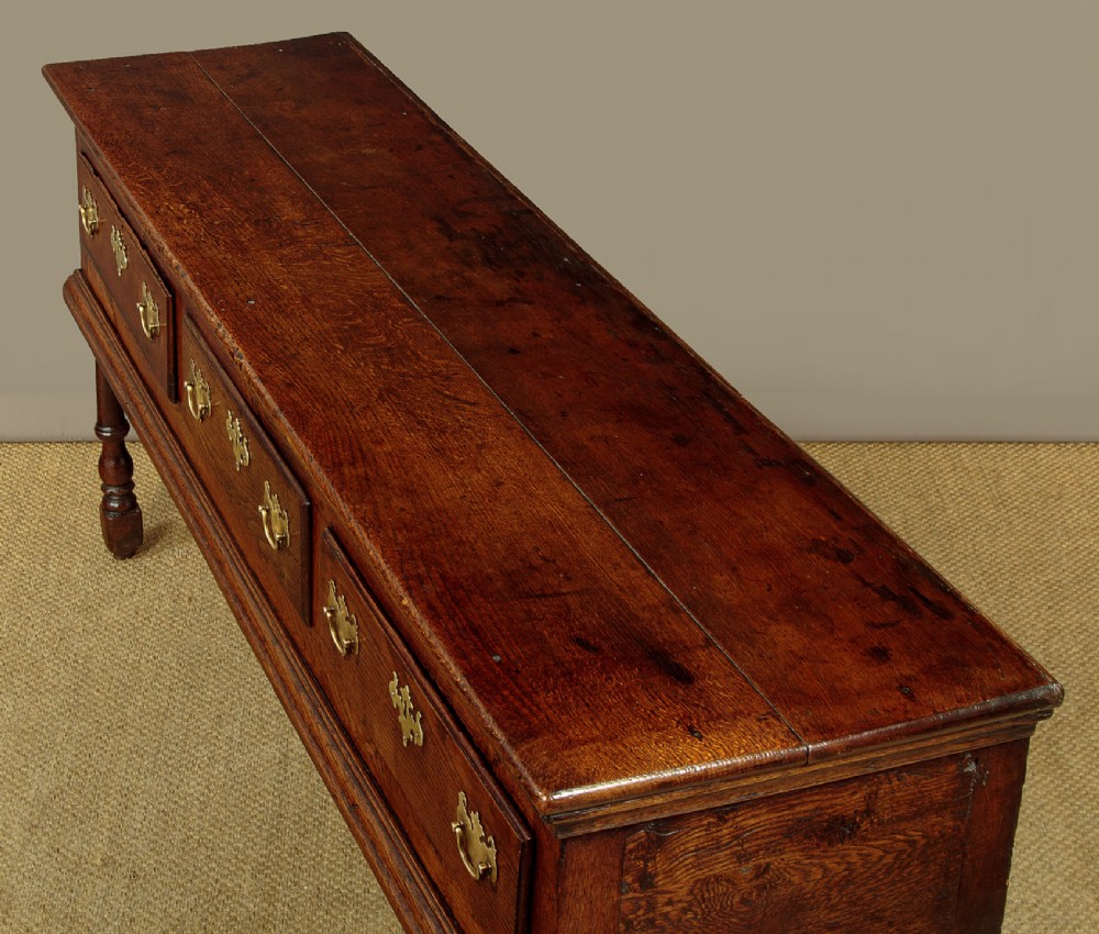 An 18th-century dark oak dresser base featuring three inline drawers with decorative brass drop handles and turned front legs. View VI