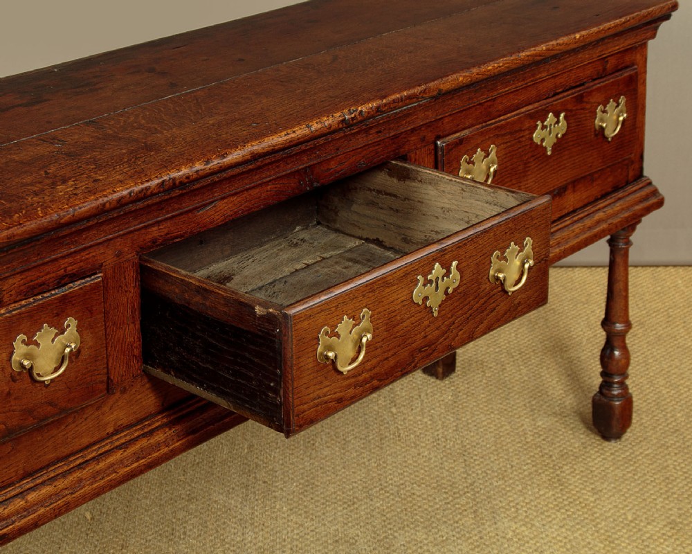 An 18th-century dark oak dresser base featuring three inline drawers with decorative brass drop handles and turned front legs. View VII