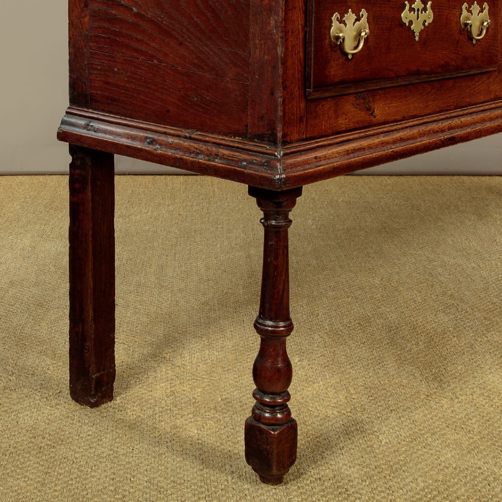 An 18th-century dark oak dresser base featuring three inline drawers with decorative brass drop handles and turned front legs. View IX