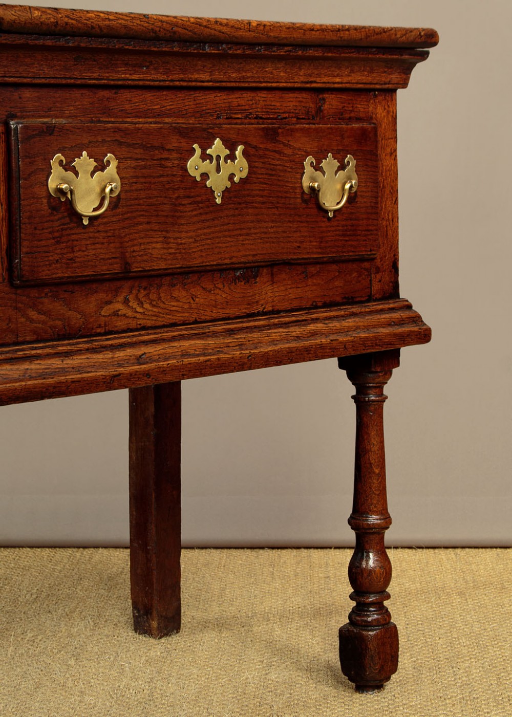 An 18th-century dark oak dresser base featuring three inline drawers with decorative brass drop handles and turned front legs. View X