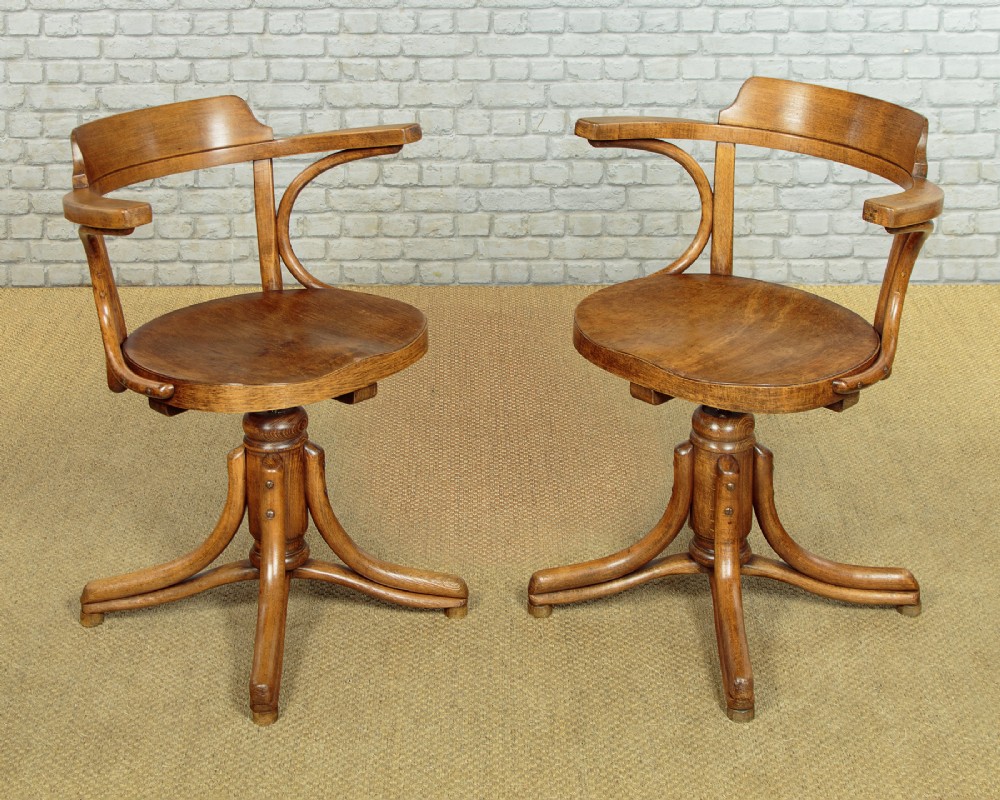 A pair of antique warm brown bentwood swivel desk chairs with curved armrests and quadruped pedestal bases.
