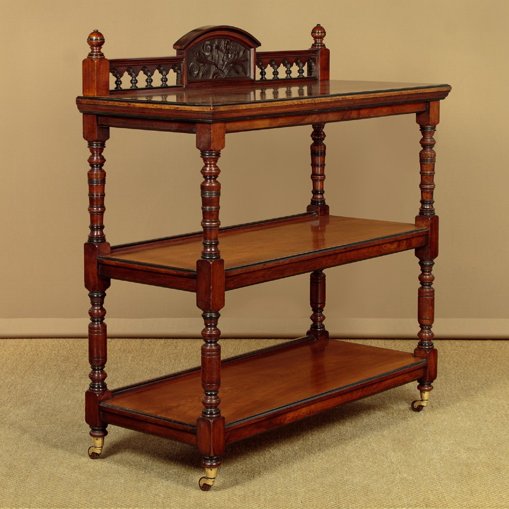 An Aesthetic Movement three-tier walnut buffet featuring turned corner columns, a carved floral back gallery, and brass castors.