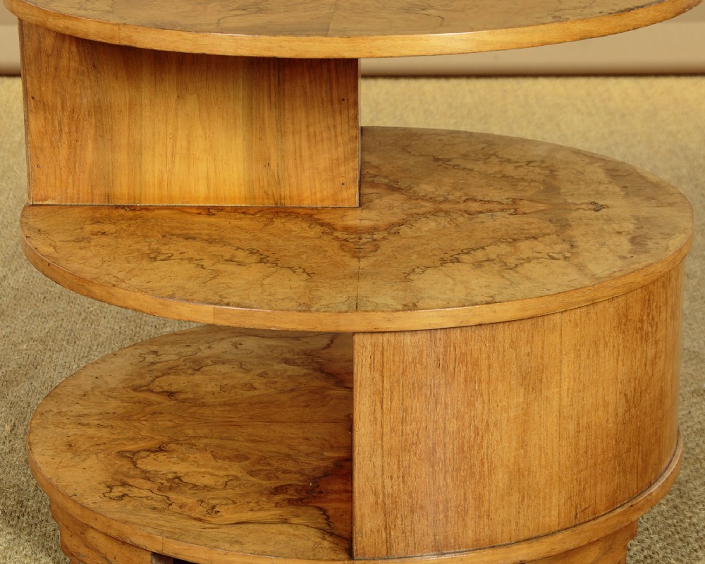 A circular three-tiered Art Deco coffee table constructed from warm golden burr timber veneer with offset geometric supports. View IX