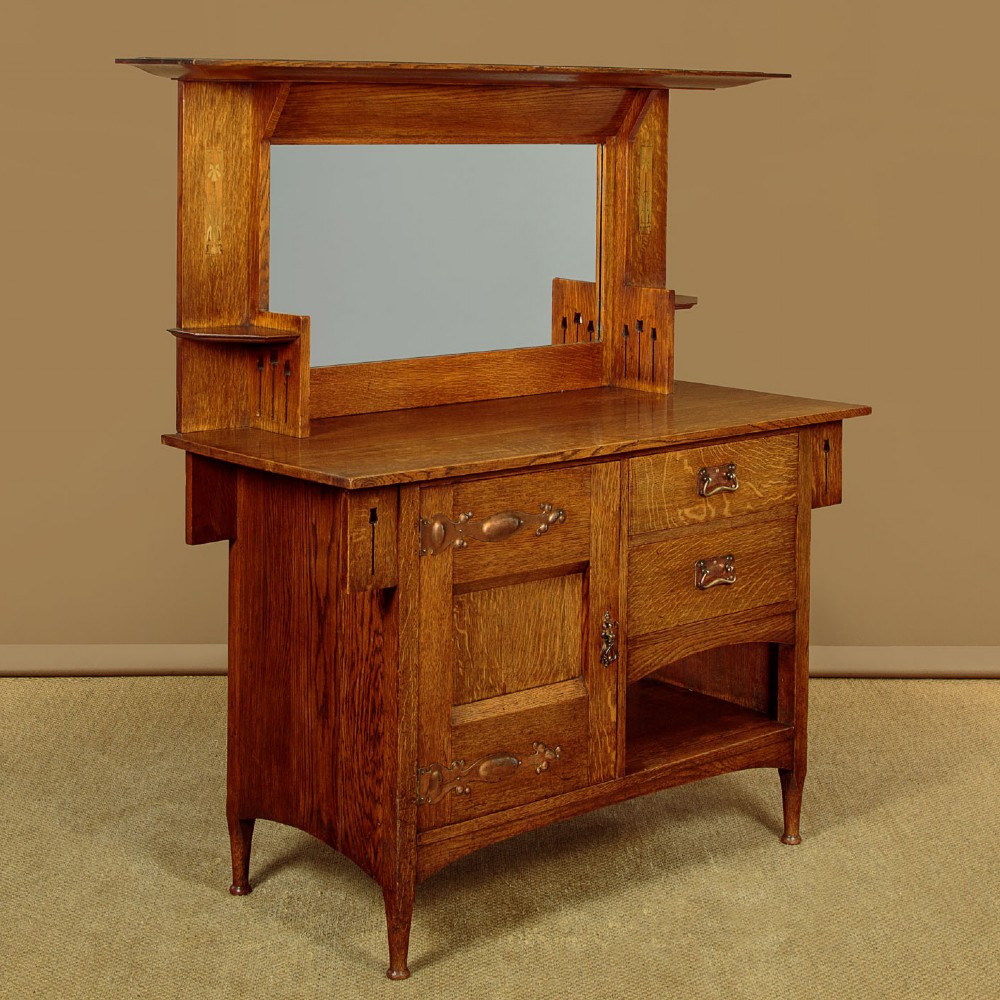 An Arts and Crafts quarter-sawn oak dresser featuring a mirrored top structure with small display shelves, inlaid floral details, hammered copper strap hinges, two drawers, and a lower cupboard.