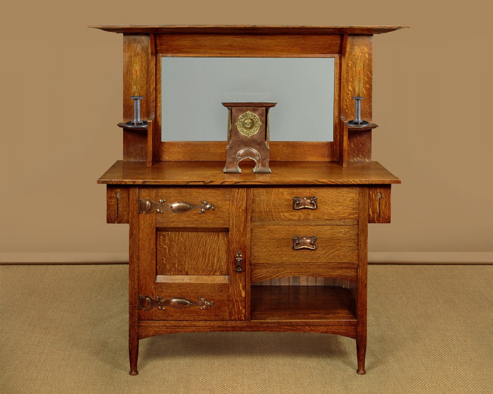 An Arts and Crafts quarter-sawn oak dresser featuring a mirrored top structure with small display shelves, inlaid floral details, hammered copper strap hinges, two drawers, and a lower cupboard. View II