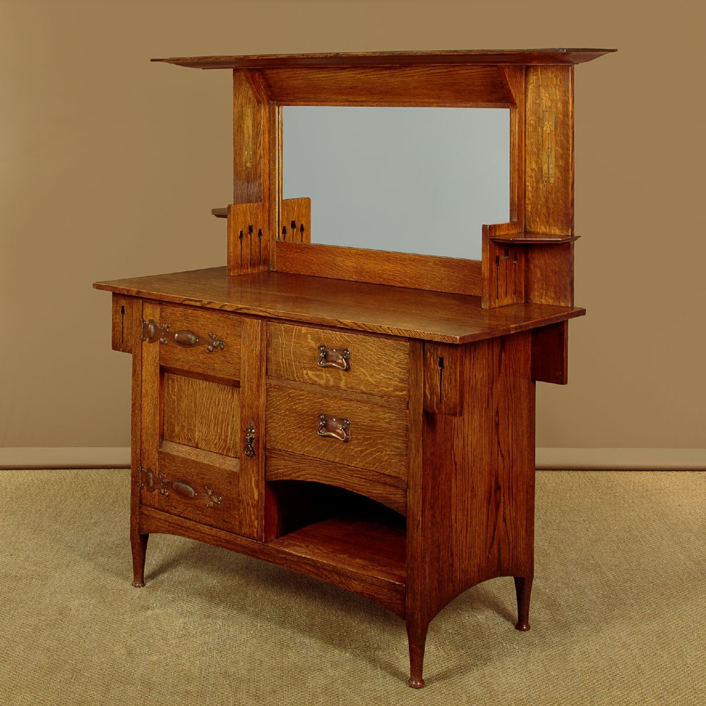 An Arts and Crafts quarter-sawn oak dresser featuring a mirrored top structure with small display shelves, inlaid floral details, hammered copper strap hinges, two drawers, and a lower cupboard. View III