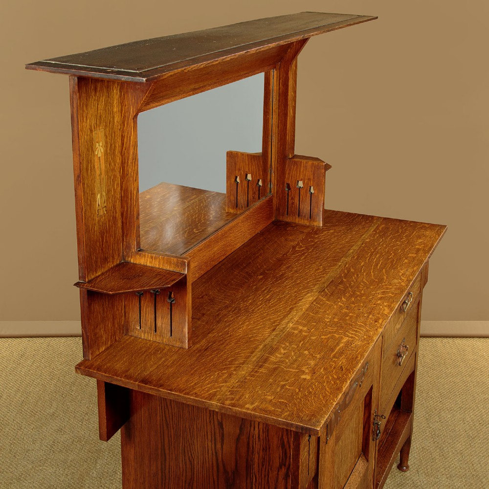 An Arts and Crafts quarter-sawn oak dresser featuring a mirrored top structure with small display shelves, inlaid floral details, hammered copper strap hinges, two drawers, and a lower cupboard. View V